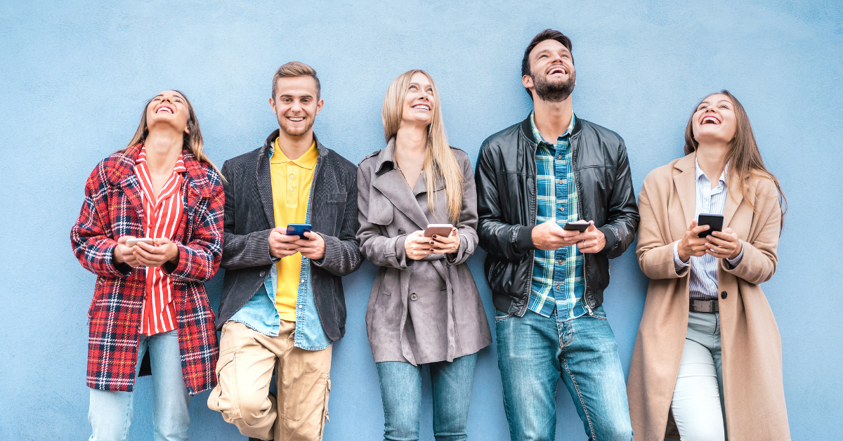 Group of 5 young adults leaning against a blue wall, all holding their phones and smiling